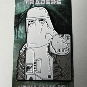 May include: A white and gray enamel pin featuring a character from Star Wars. The character is wearing a white helmet and a white hooded cloak. The pin is labeled "Dagobah Traders" and "Limited Edition Pin".