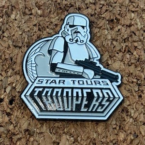 May include: A Star Tours Troopers pin featuring a Stormtrooper in white and gray. The pin has a black border and the words "STAR TOURS TROOPERS" in white lettering.