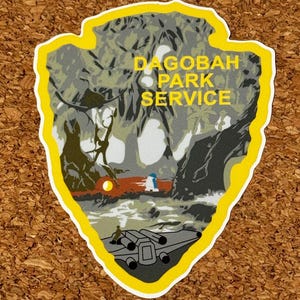 May include: A sticker with a yellow border in the shape of an arrowhead. The sticker features a forest scene with the text "Dagobah Park Service" in yellow. An X-wing fighter and a figure are visible in the design.