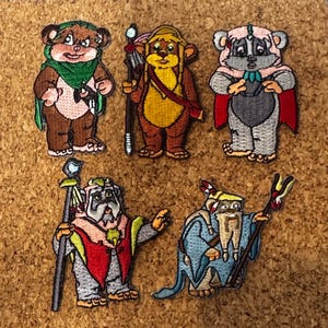 Pode incluir: Cinco patches bordados com personagens de desenhos animados. Os patches retratam várias criaturas em diferentes roupas, incluindo capas, túnicas e chapéus. A paleta de cores inclui marrom, vermelho, azul e amarelo. Cada patch tem costuras detalhadas e designs exclusivos.