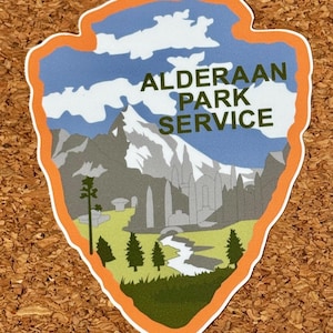 以下が含まれることがあります： 「Alderaan Park Service」のテキストが入った、矢印型のステッカー。山脈、木々、川、雲のある青い空がデザインされています。ステッカーはオレンジ色の縁取りです。