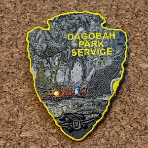 以下が含まれることがあります： 矢じり型の刺繍パッチ。黄色の「DAGOBAH PARK SERVICE」の文字が入った詳細な風景画が描かれています。パッチは黄色の縁取りで、森、水、乗り物を描いています。