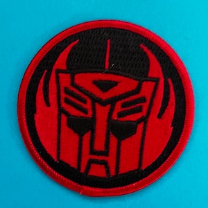 Pode incluir: Um remendo bordado vermelho e preto com um design de rosto de robô. O rosto do robô é cercado por um círculo preto com uma borda vermelha.