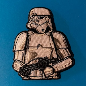 Puede incluir: Un parche bordado blanco de un Stormtrooper de Star Wars sosteniendo un rifle bláster. El parche está detallado con costuras negras y tiene un fondo blanco.