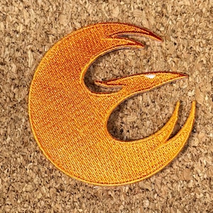 Pode incluir: Patch bordado laranja com o símbolo da Aliança Rebelde. O patch tem uma aparência texturizada e está sobre um fundo de cortiça. O design é uma forma estilizada de pássaro ou fénix.