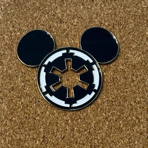 May include: A black and white enamel pin in the shape of Mickey Mouse ears. The center features a white and black Imperial symbol. The pin is set against a corkboard background.