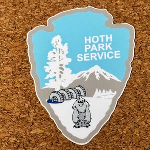以下が含まれることがあります： 薄い青色の背景に、白い文字で「HOTH PARK SERVICE」と書かれたステッカー。ステッカーには山、木、漫画のイエティが描かれています。ステッカーは矢印の形をしています。