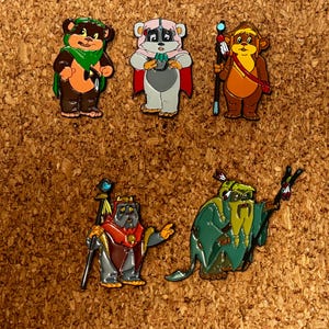 Pode incluir: Cinco pinos de esmalte coloridos com personagens de desenhos animados. Os pinos incluem um personagem semelhante a um urso com um lenço verde, um personagem rosa e cinza com uma capa vermelha e um personagem marrom com uma bengala. Os outros dois pinos são um personagem cinza com uma bengala e um personagem verde com uma bengala.