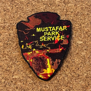 Puede incluir: Parche bordado en forma de punta de flecha, con el texto "Mustafar Park Service" en amarillo. El parche representa un paisaje de fuego en tonos rojos, naranjas y amarillos, con un borde negro.