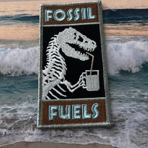 Puede incluir: Parche bordado con un esqueleto de dinosaurio bebiendo de una taza. El parche tiene las palabras "FOSSIL FUELS" en un marco rectangular. El dinosaurio es blanco, el fondo es negro y el marco es marrón con texto azul claro.