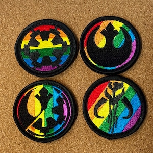 May include: Four round embroidered patches with black borders. Each patch features a symbol in the center, with a rainbow-colored design. The symbols include a gear, a bird, and a Mandalorian skull. The patches are on a brown surface.