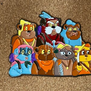 Pode incluir: Um alfinete de esmalte colorido com um grupo de personagens de desenhos animados semelhantes a ursos. Os ursos são retratados em várias cores, incluindo laranja, azul, marrom e amarelo, com expressões faciais e acessórios detalhados. O alfinete é provavelmente um item de colecionador ou uma obra de arte.