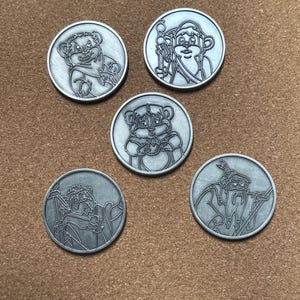 Puede incluir: Cinco monedas redondas de color plateado con diseños de personajes de dibujos animados grabados. Las monedas presentan varios personajes, incluyendo una criatura parecida a un oso y una figura con un bastón. Las monedas están dispuestas sobre una superficie marrón.