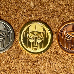 Puede incluir: Tres monedas metálicas con un diseño de rostro de robot. Las monedas son plateadas, doradas y bronce, dispuestas en fila. El fondo es una superficie texturizada y marrón.