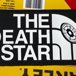 Może przedstawiać: Czarna prostokątna naszywka z białym napisem "THE DEATH STAR". Projekt zawiera grafikę Gwiazdy Śmierci i logo. Naszywka jest umieszczona na tle amerykańskiej flagi.