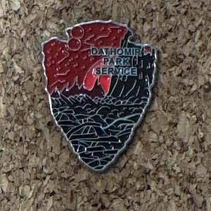 May include: A Dathomir Park Service pin in the shape of an arrowhead. The top half is red with a mountain range design, and the bottom half is black with a landscape design. The text "Dathomir Park Service" is visible.