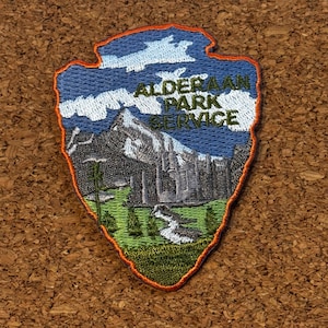 以下が含まれることがあります： 矢印型の刺繍パッチ。山岳風景が描かれ、「Alderaan Park Service」の文字が入っています。パッチには青い空、緑の草、白い川があります。
