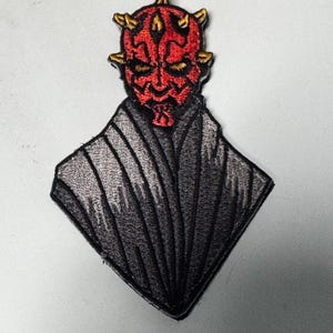 May include: Embroidered patch featuring a red-faced character with horns and black markings. The character is set against a black and gray diamond-shaped background, with detailed stitching. The patch is likely for clothing or accessories.