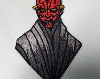 Può includere: Toppa ricamata raffigurante un personaggio con il volto rosso con corna e segni neri. Il personaggio è posto su uno sfondo a forma di diamante nero e grigio, con cuciture dettagliate. La toppa è destinata a vestiti o accessori.