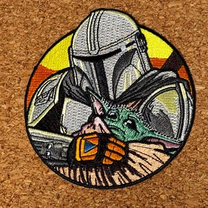 May include: Embroidered patch featuring The Mandalorian and Grogu from the Star Wars series. The patch is circular, with a detailed depiction of the characters in shades of gray, green, orange, and yellow, set against a sunset background.