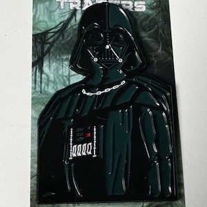 May include: A black and green enamel pin featuring Darth Vader from Star Wars. The pin is a limited edition and has the text "Dagobah Traders" and "Limited Edition Pin" on the card.