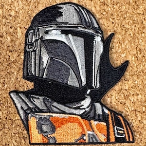 May include: Embroidered patch featuring the helmet and part of the armor of a character from a science fiction series. The patch is primarily black, gray, and orange, with detailed stitching. The design is a stylized representation of the character's helmet and shoulder armor.