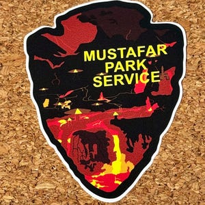 May include: A sticker with a black background and a red and yellow landscape design. The text "MUSTAFAR PARK SERVICE" is in yellow. The sticker is shaped like an arrowhead.