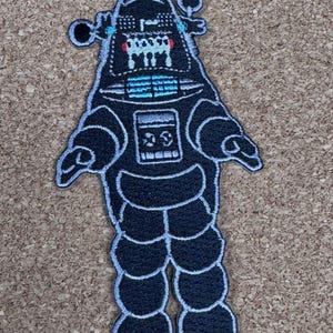 May include: Black robot patch with white stitching. The robot has a detailed design with a head, body, arms, and legs. The robot's head has two small antennas and a detailed face. The patch is on a tan background.