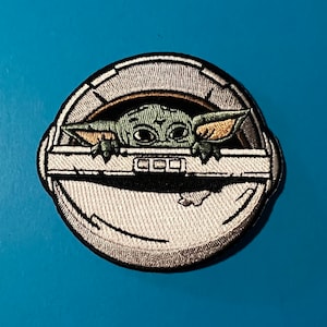 Puede incluir: Un parche bordado blanco y gris con un personaje alienígena verde asomándose desde una cápsula redonda. El alienígena tiene orejas grandes y ojos grandes. La cápsula tiene un borde negro y un interior blanco.