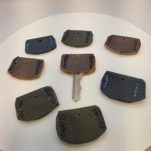 May include: A collection of handmade leather key covers in various colors, including brown, black, and green. Each cover is stitched with contrasting thread and features a small hole for a key ring. One key is inserted into a cover.