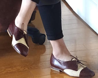 May include: Brown leather shoes with a beige woven detail on the toe and laces. The shoes have a pointed toe and a low heel.