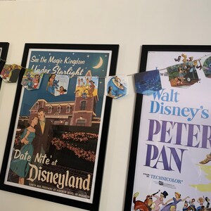 May include: A framed poster for Walt Disney's Peter Pan movie with a string of Disney-themed paper flags hanging above it. The flags feature characters like Stitch, Cinderella, and Ariel. The poster is in a black frame and has a white background with the title "Walt Disney's Peter Pan" in large, bold letters. The poster also features a color by Technicolor logo.