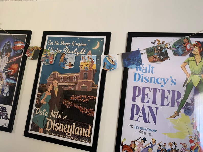 May include: A framed poster for Walt Disney's Peter Pan movie with a string of Disney-themed paper flags hanging above it. The flags feature characters like Stitch, Cinderella, and Ariel. The poster is in a black frame and has a white background with the title "Walt Disney's Peter Pan" in large, bold letters. The poster also features a color by Technicolor logo.