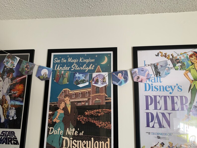 May include: A banner with images of Disney characters hangs between two framed posters. The poster on the left features a Star Wars scene. The poster on the right is for the movie Peter Pan and features the text "Walt Disney's Peter Pan" and "Color by Technicolor". The banner features images of characters from Disney movies such as Cinderella, Frozen, and The Little Mermaid.