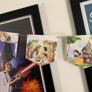 May include: A string of pennant flags with illustrations from the fairy tale Snow White. The flags feature Snow White, the Evil Queen, and the Prince. The flags are hung on a wall with a framed poster in the background.
