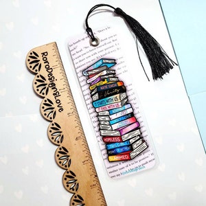 K&ouml;nnte beinhalten: Ein transparentes Acryl-Lesezeichen mit einem Stapel bunter B&uuml;cher, die darauf gedruckt sind. Die Buchtitel beinhalten "Maybe Someday", "Verity", "It Starts With Us", "It Ends With Us", "Ugly Love", "Gay Love", "Hopeless", "This Girl", "Point of Retreat" und "Slammed". Das Lesezeichen hat einen schwarzen Quasten.
