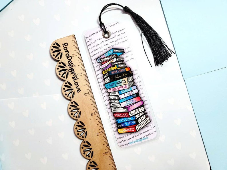 K&ouml;nnte beinhalten: Ein transparentes Acryl-Lesezeichen mit einem Stapel bunter B&uuml;cher, die darauf gedruckt sind. Die Buchtitel beinhalten "Maybe Someday", "Verity", "It Starts With Us", "It Ends With Us", "Ugly Love", "Gay Love", "Hopeless", "This Girl", "Point of Retreat" und "Slammed". Das Lesezeichen hat einen schwarzen Quasten.