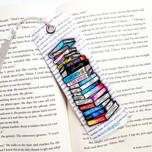 K&ouml;nnte beinhalten: Ein farbenfrohes Lesezeichen mit einem Stapel B&uuml;cher mit Titeln wie "It Starts With Us", "Ugly Love", "Hopeless" und "This Girl". Das Lesezeichen ist mit einem wei&szlig;en Quasten verziert.