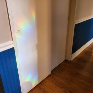 May include: Interior shot of a hallway with a white door and blue wainscoting. The walls are painted in a light beige color. A rainbow of light is cast on the door. The floor is made of wood.