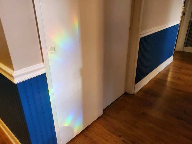 May include: Interior shot of a hallway with a white door and blue wainscoting. The walls are painted in a light beige color. A rainbow of light is cast on the door. The floor is made of wood.