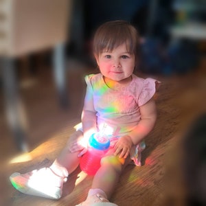 May include: A young child sits on a wooden floor, illuminated by a rainbow of light. The child wears a pink shirt with the text "Roro Design" and a matching skirt. A red and blue sippy cup rests in their lap, and they wear white sneakers.