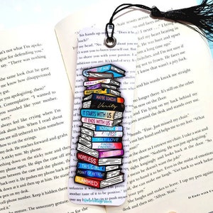 K&ouml;nnte beinhalten: Ein farbenfrohes Lesezeichen mit einem Stapel Emaille-Pins, die Buchcover darstellen. Die Pins sind in einem Stapel angeordnet, wobei die Titel der B&uuml;cher sichtbar sind. Das Lesezeichen ist an einem schwarzen Quasten befestigt.
