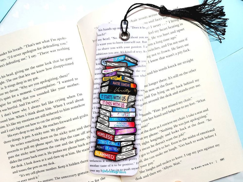 K&ouml;nnte beinhalten: Ein farbenfrohes Lesezeichen mit einem Stapel Emaille-Pins, die Buchcover darstellen. Die Pins sind in einem Stapel angeordnet, wobei die Titel der B&uuml;cher sichtbar sind. Das Lesezeichen ist an einem schwarzen Quasten befestigt.
