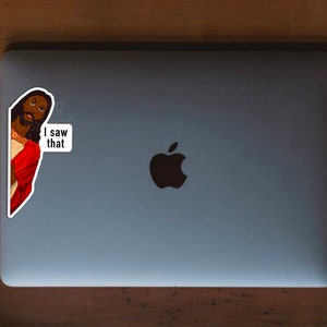 Pode incluir: Um adesivo de uma pessoa com pele escura e cabelo comprido vestindo uma t&uacute;nica vermelha e olhando por cima do ombro. O adesivo est&aacute; em um laptop prateado com um logotipo da Apple preto. O adesivo diz "Eu vi isso".