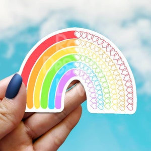 May include: A rainbow-colored sticker with a design of hearts forming an arch. The sticker is held in a hand against a blue sky background.