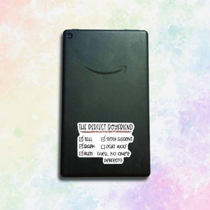 May include: A black tablet with a white sticker that says "THE PERFECT BOYFRIEND" with a checklist of qualities. The checklist includes "Tall", "Sexy Accent", "Dark", "Port Wolf", and "Rich (Well, no one's perfect!)"