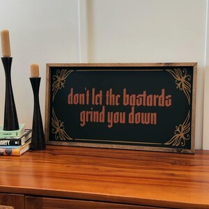 May include: A rectangular framed sign with the text "don't let the bastards grind you down" in a copper colour on a black background. The frame is wood-toned, and the sign has decorative corner accents.