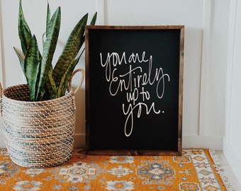 May include: A black framed sign with white calligraphy that reads "You are entirely up to you." The sign is sitting on a brown wooden floor with an orange and blue patterned rug in front of it. A potted snake plant is in the background.