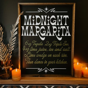 May include: A black framed chalkboard sign with white text that reads "MIDNIGHT MARGARITA" and instructions for making a margarita. The sign is on a shelf with a brown wood surface and a black and white tiled floor.