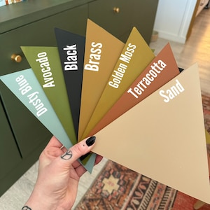 Op de afbeelding: Een waaier met acht verfstalen. De kleuren zijn Stofblauw, Avocado, Zwart, Messing, Gouden Mos, Terracotta, Zand en een donkergroen.